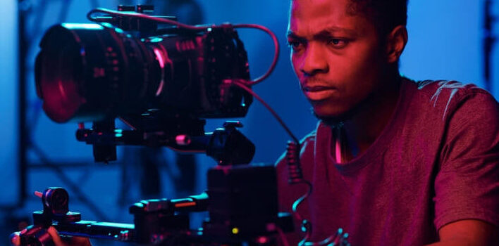 Man operating a professional film camera on a movie set with colorful lighting.